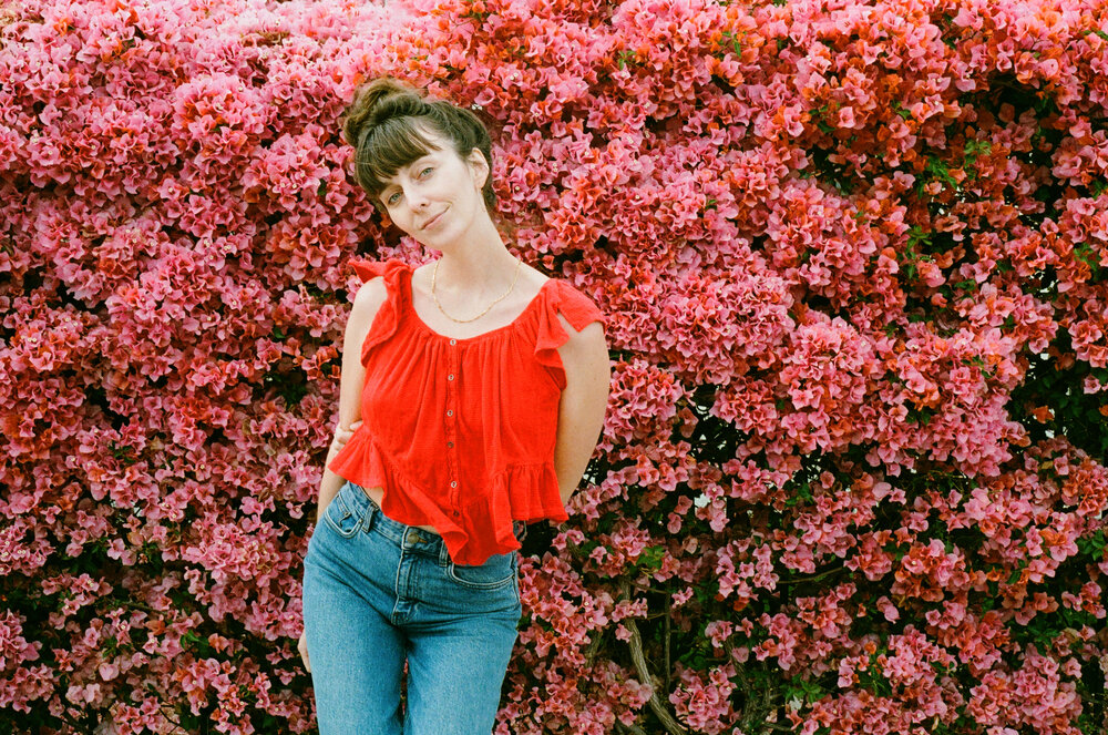 Anna Ash Red Shirt Blue Jeans Standing in Front of Pink Flowers