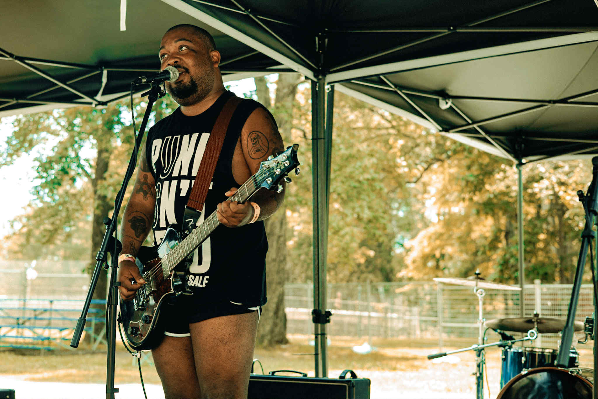 musician black guy fawkes playing guitar and singing into a microphone at an outside festival.